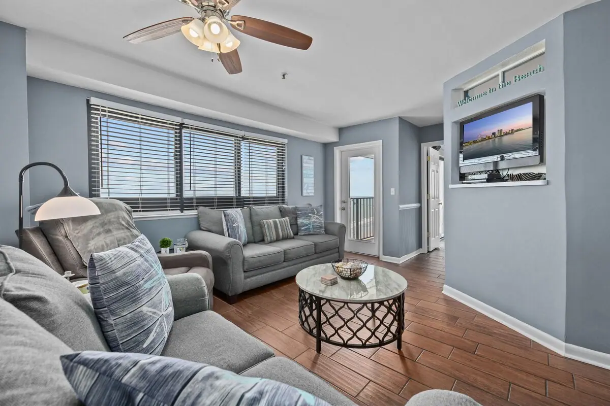 Living room with gray sofas, wide windows, wood flooring, and a wall-mounted television