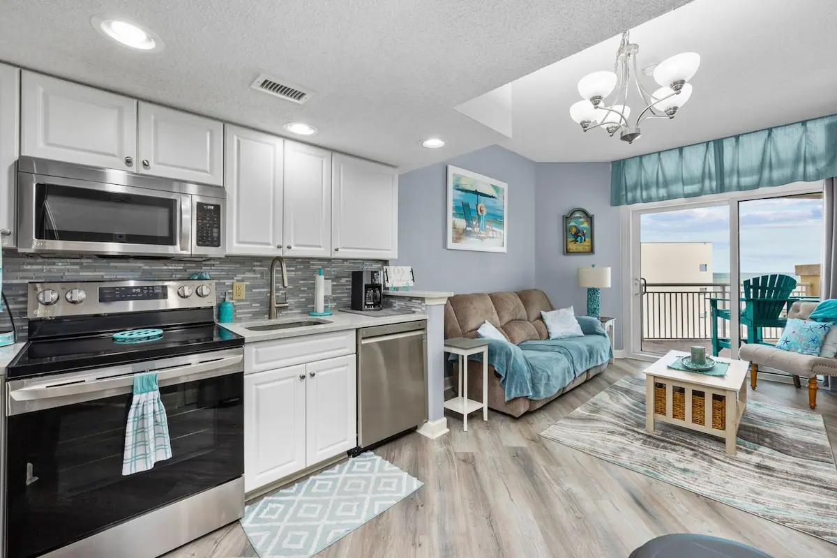 White kitchen with dark appliances opening to a living room with balcony doors