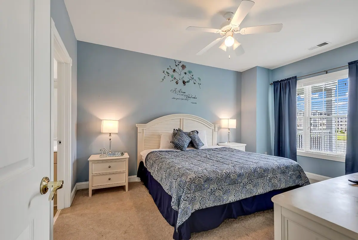 Blue-walled bedroom with a patterned blue bedspread, white headboard, and window