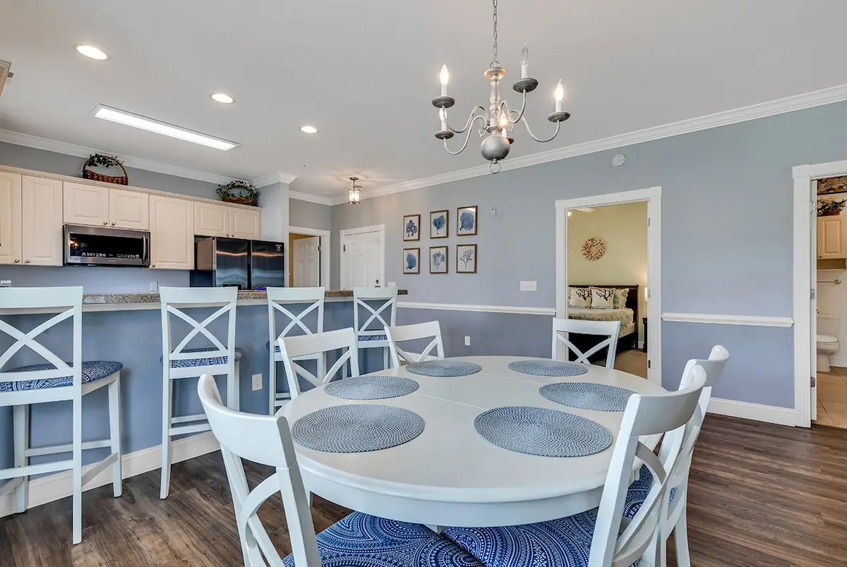 Round white dining table beneath a chandelier, with the kitchen behind it