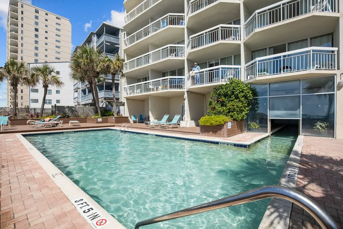 Outdoor pool beside a building with stacked balconies and palm trees