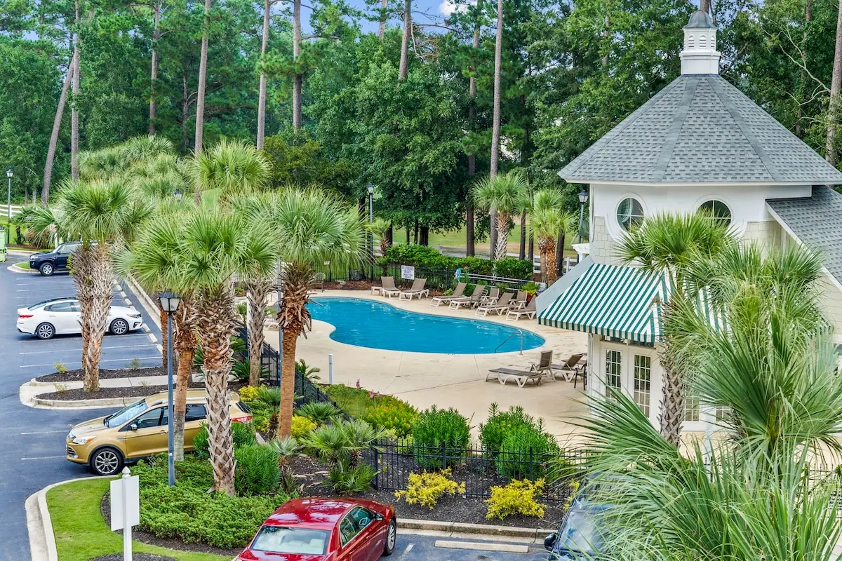 Outdoor pool among palms beside a clubhouse and parking area